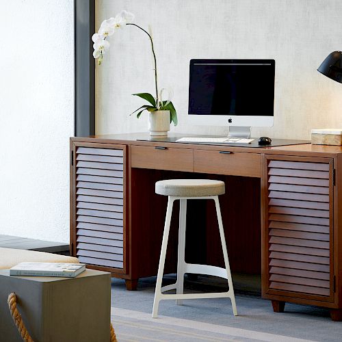 A modern desk setup with a computer, lamp, plant, and a stool in a well-organized, stylish office space. A window with blinds is on the left.