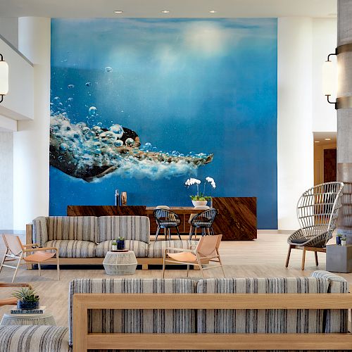 A modern lobby with striped sofas, chairs, a front desk, and a large wall mural of an underwater scene with a swimming sea turtle, and decorative plants.