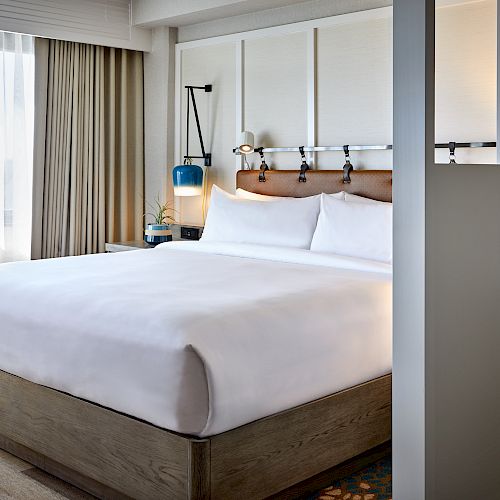 A modern hotel room with a large bed, bedside lamp, and window with curtains, featuring stylish decor and ample natural light.