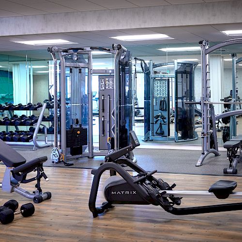 The image shows a gym with various exercise equipment, including a rowing machine, a weight bench, free weights, and multiple weight machines.