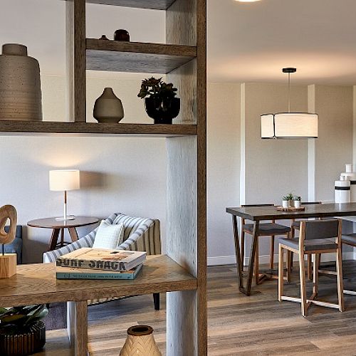 The image shows a modern living and dining area with stylish wooden furniture, a bookshelf, a lamp, and decorative items on display.