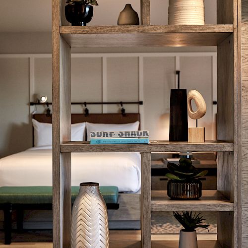 The image shows a cozy bedroom with a bed and decor, featuring a wooden shelving unit displaying various vases and items, separating the space.