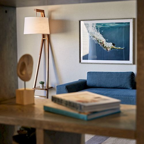 The image shows a cozy living room with a blue sofa, a wooden floor lamp with a white shade, and a framed wall art. Books are on a shelf.