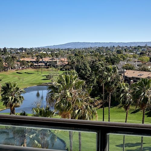 The image shows a scenic view of a golf course with a pond, surrounded by palm trees and residential houses, taken from a balcony railing.