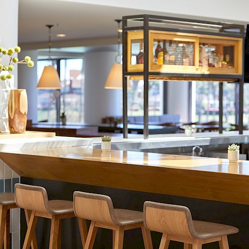 A modern bar area with wooden stools, a countertop, decorative plants, and a TV, in a well-lit room.