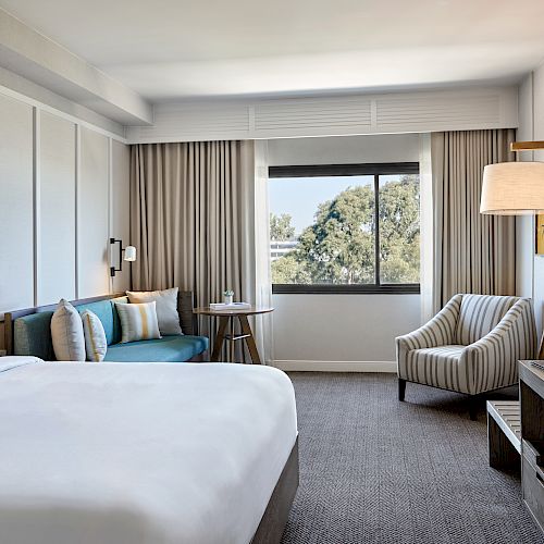 A modern hotel room with a bed, couch, armchair, desk, TV, floor lamp, and a large window with curtains.