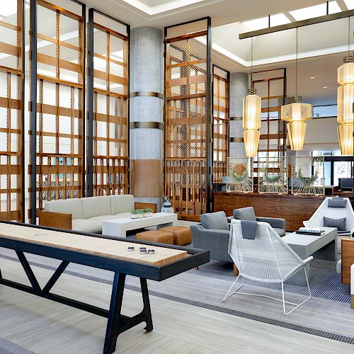 The image shows a modern lounge area with stylish seating, a shuffleboard table, and hanging lights, featuring wooden and metal accents.