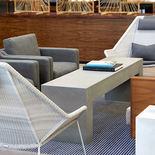 The image shows a modern seating area with gray chairs, a concrete table, a small white table with a plant, and books on the main table.