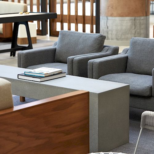 The image shows a modern lounge area with gray armchairs, a wooden bench with a cushioned backrest, a gray rectangular table with books on it, and other seating.