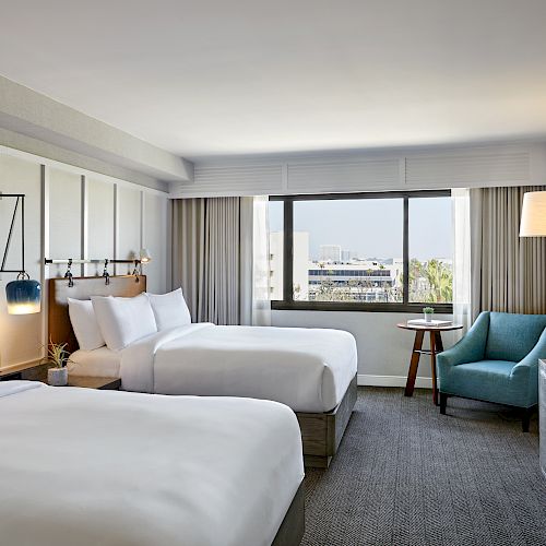 This image shows a modern hotel room with two beds, a window with a view, a turquoise armchair, a wooden table, and a TV on a cabinet.