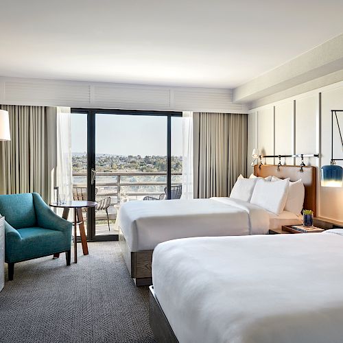 A modern hotel room with two large beds, a teal armchair, a TV, a lamp, and a balcony with a city view. Neutral tones dominate the decor.