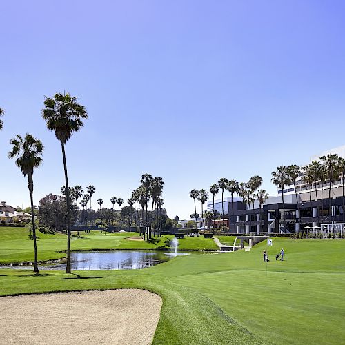 A sunny golf course with palm trees, a sand bunker, a pond, and buildings in the background. Two people are seen walking on the green.