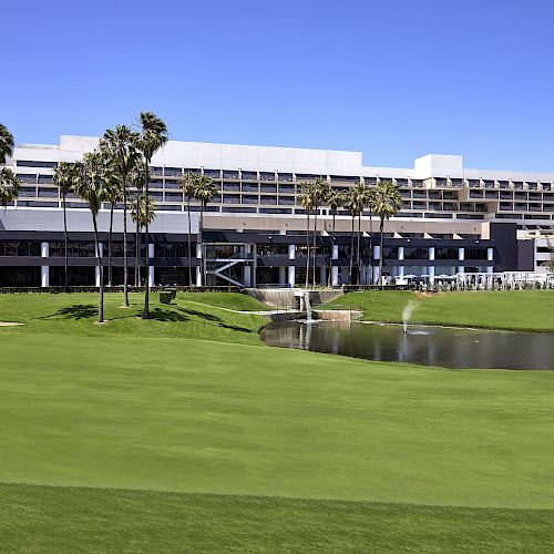 This image shows a modern building with multiple floors, a landscaped green lawn, a pond, and several palm trees under a clear blue sky.