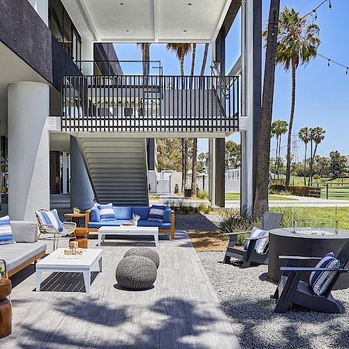 An outdoor patio with modern seating, a fire pit, and string lights beside a large building, over a green lawn with scattered palm trees.