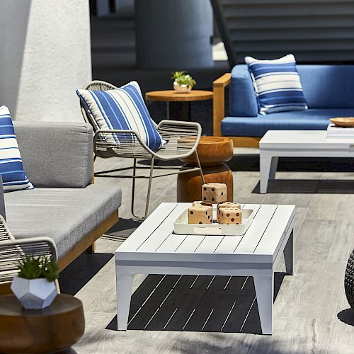 An outdoor seating area with gray and blue furniture, striped cushions, wooden side tables, and a white coffee table with decor, set on a wooden deck.