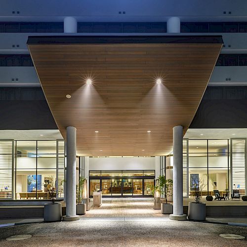 The image shows the entrance of a modern building at night, with a wooden canopy, large glass windows, and illuminated interiors.