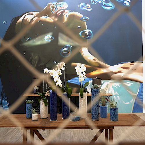 A table with various vases and flowers is in front of a large underwater mural, viewed through a grid-like barrier.