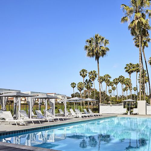 A poolside area with sun loungers under canopy beds, surrounded by tall palm trees and a clear, blue sky in the background.