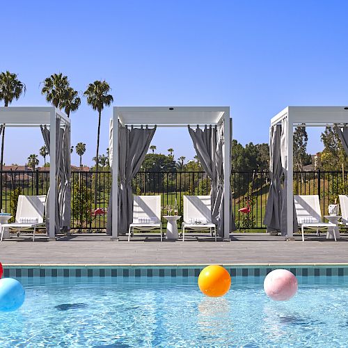 A poolside scene with three cabanas, several lounge chairs, and colorful beach balls in the water, surrounded by palm trees under a clear blue sky.