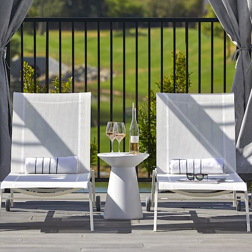 Two lounge chairs sit under a canopy, with a small table and wine glasses between them; a scenic view of a green landscape in the background.