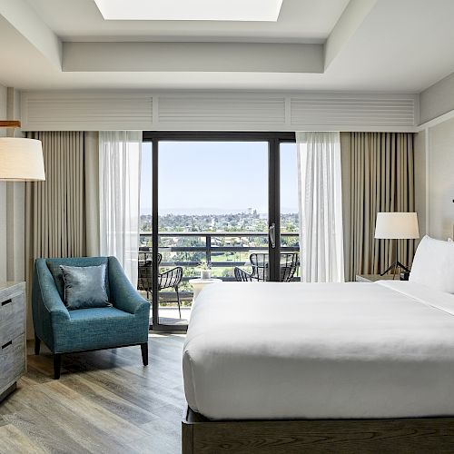 A modern hotel room features a bed, television, armchair, and lamps with a balcony view of the outdoors, blending comfort and style perfectly.