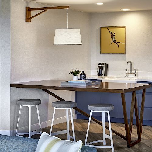 A modern kitchenette with a wooden table, three stools, a small sink area, a coffee maker, a plant, books, and a picture on the wall.