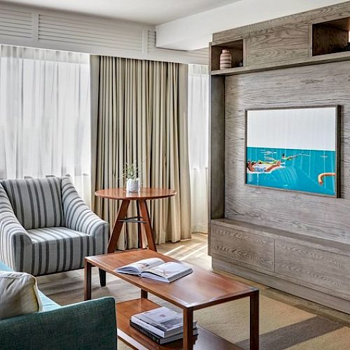 The image shows a modern living room with a teal sofa, striped armchair, coffee table with books, large window, and wooden shelving with decor.