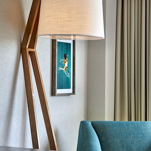 The image shows a modern living space with a wooden floor lamp, a framed picture, a teal armchair, and closed curtains in the background.