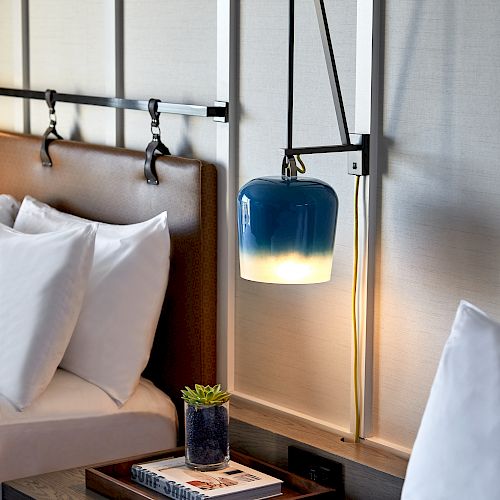 The image shows a section of a neatly made bed with white linens, a bedside table with books and a small plant, and a blue hanging lamp.