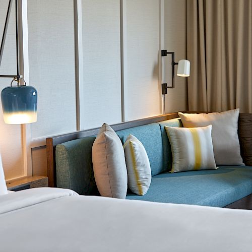 This image shows a cozy hotel room with a bed, a teal sofa adorned with pillows, wall lamps, and a small round table with a plant and books.