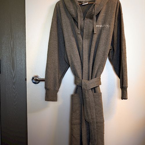 The image shows a gray bathrobe hanging on a wooden hanger against a white door. The robe has a waist tie and long sleeves.