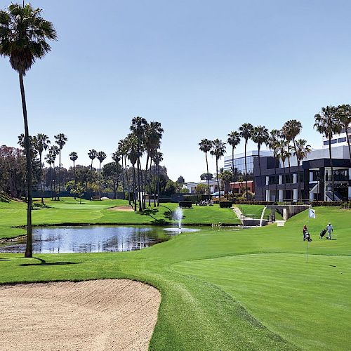 A picturesque golf course with palm trees, a pond, a sand bunker, and a modern building in the background on a bright, sunny day.