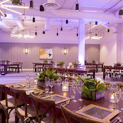 A modern banquet hall is set up with wooden tables, chairs, glassware, floral centerpieces, ambient lighting, and contemporary ceiling fixtures.