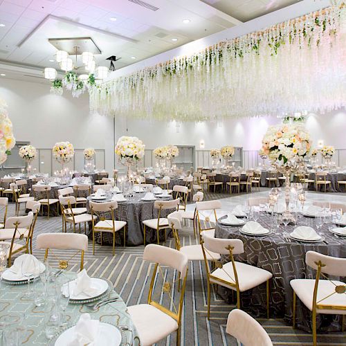 A beautifully decorated event hall with round tables covered with grey tablecloths, elegant floral centerpieces, and white chairs. There is a white ceiling.