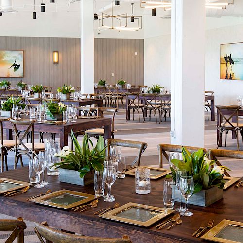 An elegantly decorated banquet hall featuring tables set with glassware, cutlery, and potted plants; modern wall art and overhead lighting fixtures complement the setting.