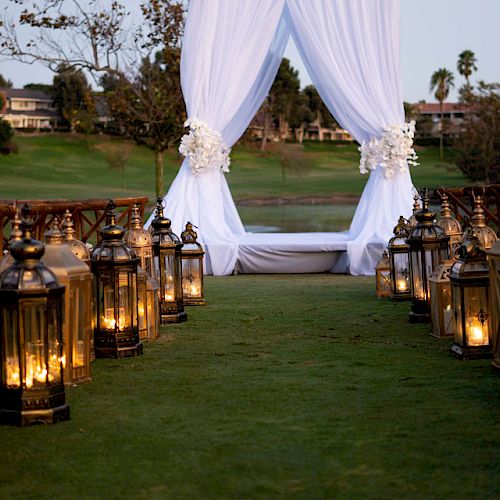 An outdoor wedding setup features an aisle lined with lanterns, leading to a white draped arch with floral decorations, set against a green landscape.