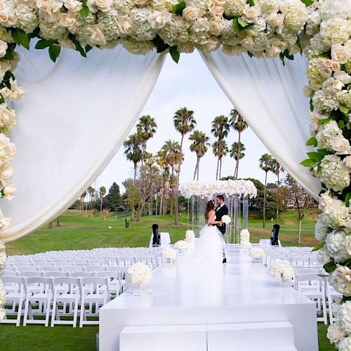 The image shows a wedding ceremony set up outdoors with a floral arch, white chairs, and palm trees in the background.