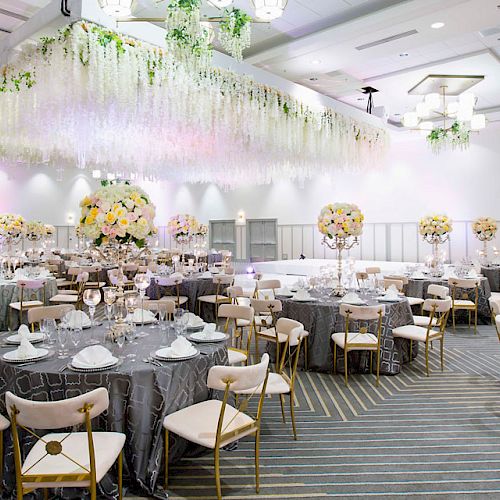 The image shows an elegantly decorated banquet hall with round tables, floral centerpieces, and a canopy of hanging flowers, ready for a formal event.