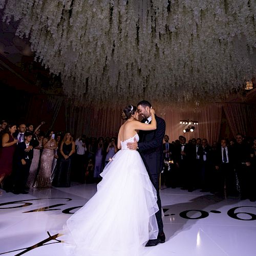 A couple is dancing in the center of a large, elegantly decorated room with a crowd of people watching and taking photos, likely at a wedding.