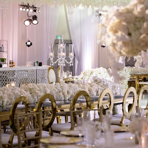 This image shows an elegant wedding reception setup with white floral arrangements, gold chairs, a bar in the background, and decorative lighting.