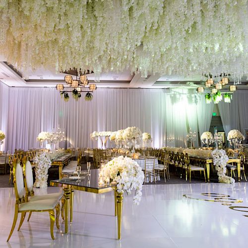 An elegantly decorated wedding reception venue with white floral arrangements, chandeliers, a glossy white dance floor, and draped walls at the sentence.