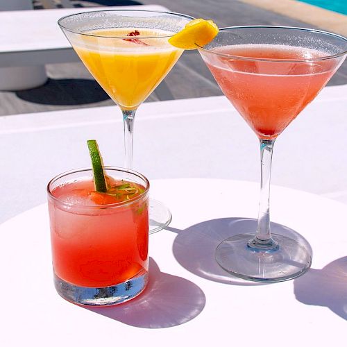 Three cocktails are on a white round table: two in martini glasses and one in a rocks glass, against a backdrop of a pool.