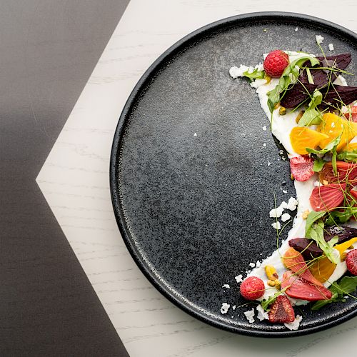 A black plate is artfully arranged with vibrant salad ingredients, including various greens, raspberries, citrus slices, and beets, garnished with microgreens.