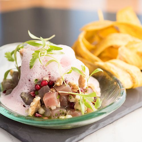 The image shows a gourmet dish with diced tuna, garnished with greens and foam, accompanied by a side of plantain chips on a slate plate.