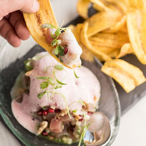 The image shows a plate of food with plantain chips. Someone is scooping up a portion with a chip, while another hand holds a spoon.