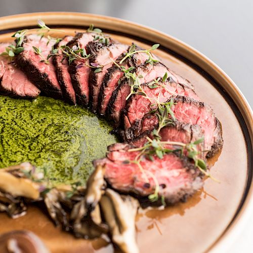 The image shows a plated dish of sliced steak served with green sauce, mushrooms, and garnished with fresh herbs on a brown plate.