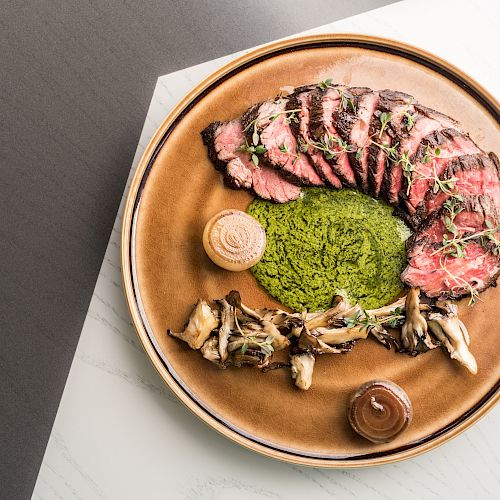 This image shows a gourmet dish with sliced beef, a green puree, mushrooms, and garnishes on a brown plate placed on a light-colored surface.