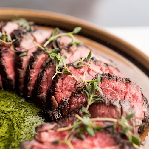 The image shows a beautifully arranged plate of sliced, medium-rare steak garnished with herbs, accompanied by a green sauce.