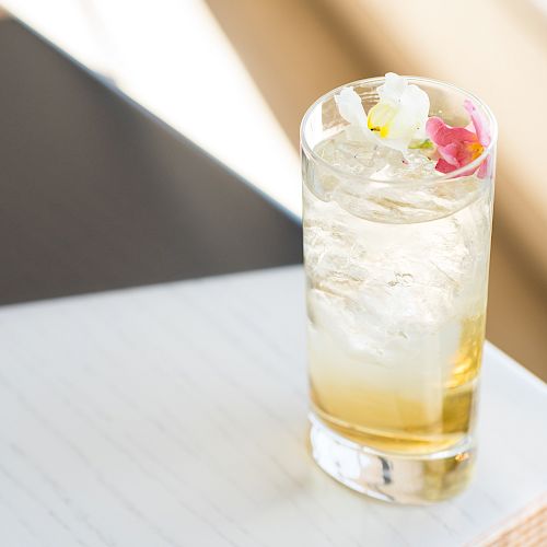 A tall glass filled with a cold drink, ice cubes, and garnished with flowers, placed on a light-colored surface.