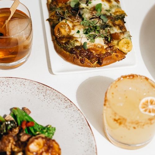 A table set with a flatbread pizza, a salad, a cocktail with lime, and a glass with an amber drink and a wooden stirrer ends the sentence.